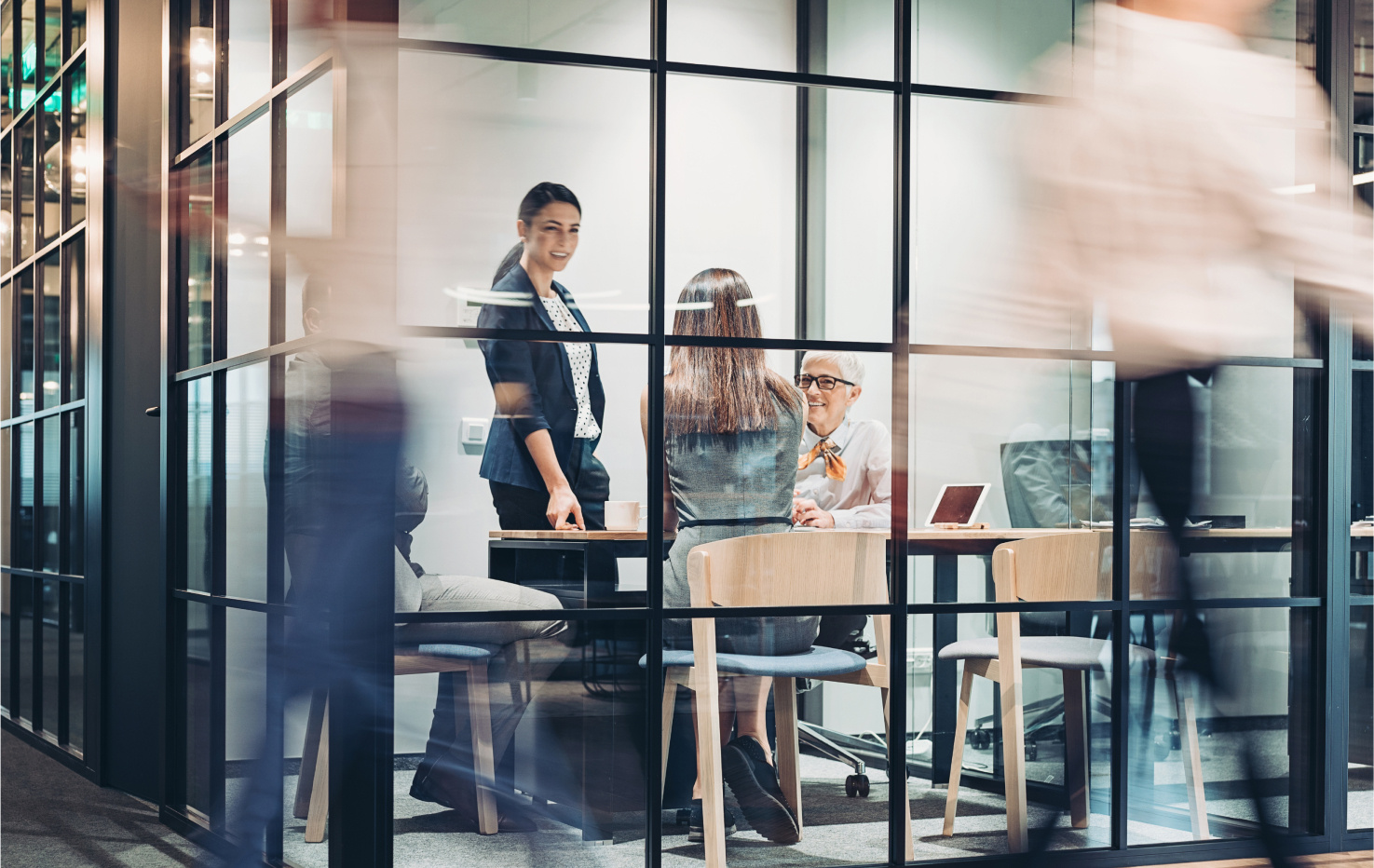 Group of professionals meeting in a glass-walled office with people walking by in the foreground.